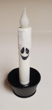 A white ghost-shaped LED candle with a black smiling face, displayed in a black candle holder against a plain beige background.