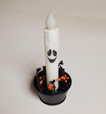 A Halloween-themed decorative candle in a black cup. The candle is white with a spooky black face, and the base of the cup features small black bats and orange pumpkin accents.