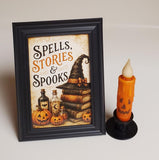 A framed Halloween-themed art print with the text 'SPELLS, STORIES & SPOOKS' beside an orange jack-o'-lantern candle holder, set against a plain light beige background.