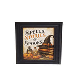 A framed Halloween-themed art print with the text 'Spells, Stories & Spooks' above illustrations of stacked books, a witch hat, potion bottles, and jack-o'-lanterns, shown in a black frame on a light beige background.